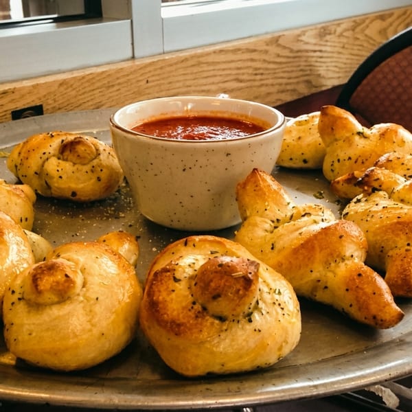 Garlic knots with herbs and spices on a metal tray, served with a side of marinara sauce.