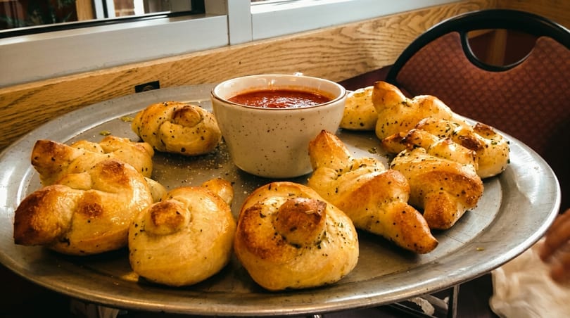 Garlic knots with herbs and spices on a metal tray, served with a side of marinara sauce.